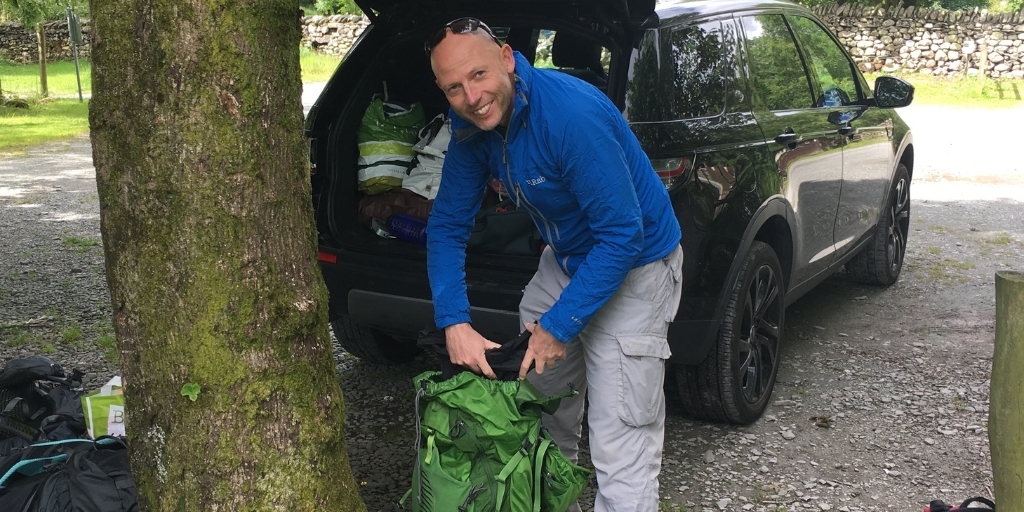 Andy crouches behind a black vehicle, sorting out a backpack for walking