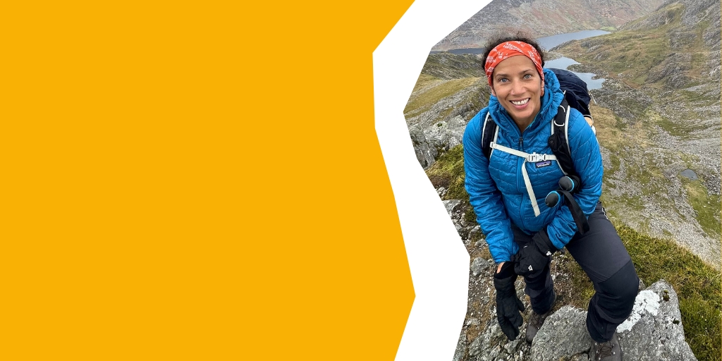 Carla smiling to camera on top of a hilly peak, overlooking a valley 