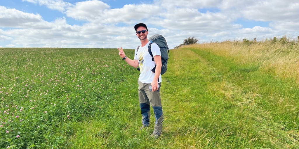 Gavin smiling to camera and posing with a peace sign on his hand, in the middle of a lush green meadow on a sunny summers' day