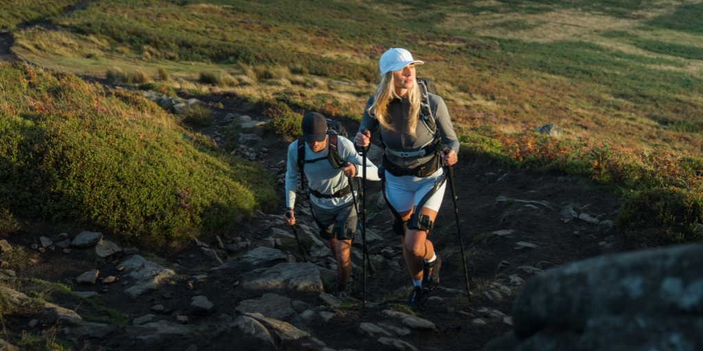 A young man and young woman are hiking up a steep, scrambly footpath. They are wearing hiking gear and using hiking sticks because of the rocky terrain. Either side of the path is green and orange moorland. It's a sunny day and the sun is low in the sky and facing towards the man and woman, the woman is squinting as she looks across the landscape .