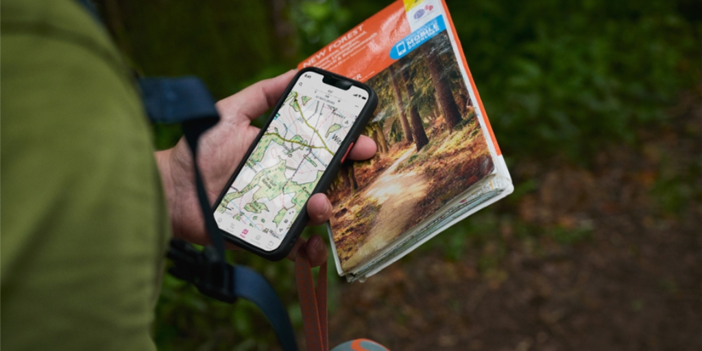 A close-up view of an individual holding a smartphone in one hand and a folded paper map in the other while standing on a forest path. The smartphone screen displays a detailed topographic-style map with footpaths marked in green. The paper map shows a woodland scene on the cover. The individual is wearing an outdoor backpack with a visible shoulder strap. The background is blurred with dense green foliage, suggesting a woodland environment.