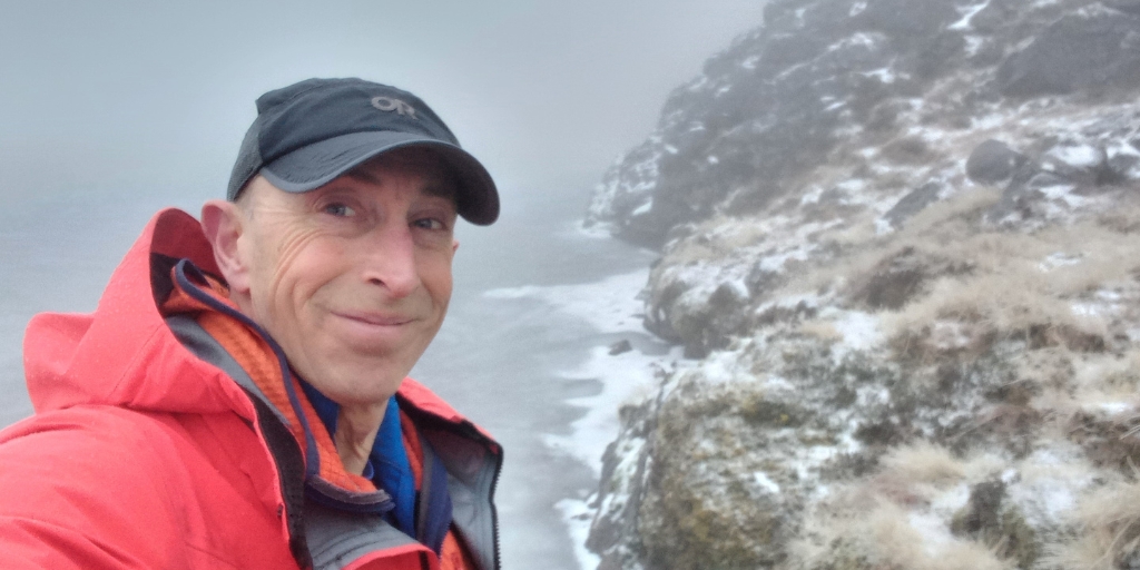 Craig is wearing a red outdoor jacket and a black cap is standing on a snowy, rocky hillside. He is holding the camera at arm’s length to take a self‑portrait. Behind him, the landscape shows a steep slope covered in patches of snow, rocks, and sparse vegetation. Mist or low cloud obscures much of the background, creating a foggy atmosphere. The ground appears icy near the edge of a frozen or partially frozen body of water.