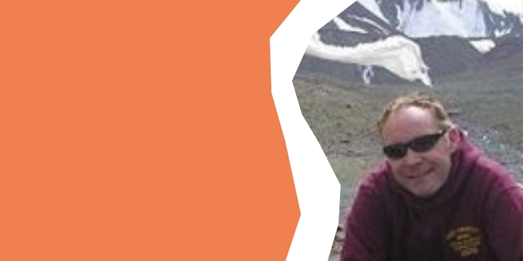 Steve is smiling to camera in a mountainous region, wearring sunglasses