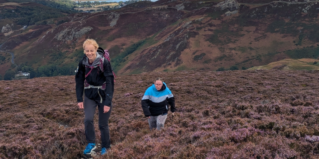 Two hikers are walking uphill through a wide expanse of purple heather on open moorland. The hiker in front is wearing a dark outdoor jacket, fitted trousers and blue walking shoes, and is carrying a backpack with straps across the front. The second hiker, slightly behind and to the right, is wearing a colour‑blocked jacket in shades of black, white and blue, along with patterned trousers. The terrain is covered in dense, low heather with earthy patches between the plants. Behind the hikers, steep hills and ridges stretch across the landscape, marked with rocky outcrops and a mixture of brown, green and purple tones. A valley and distant fields are visible further back under a cloudy sky.