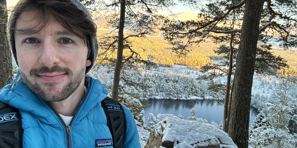 Selfie of Chris, wearing a light blue insulated jacket and carrying a backpack, standing among tall pine trees in a snowy forest. The ground and rocks are covered in snow, and a frozen or partially frozen lake lies below the viewpoint. Sunlight illuminates the distant forest, creating a bright contrast with the shaded foreground. Chris is smiling to camera in the foreground.
