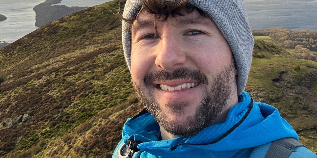 A selfie of Chris, wearing a bright blue outdoor jacket and carrying a backpack, standing on a hill overlooking a wide, scenic landscape. Below, rolling green hills lead toward a body of water dotted with small islands. The sky is filled with soft clouds and warm light, giving the scene a peaceful, expansive feel. Distant mountains frame the horizon. Chris smiles to camera in the foreground.