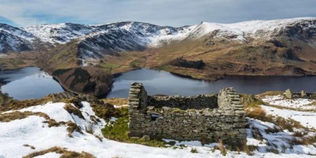 View of a hilly valley on a sunny winter day. Snow sits at the peaks while sunlight reflects if the reservoir. In the foreground, is an old stone ruin