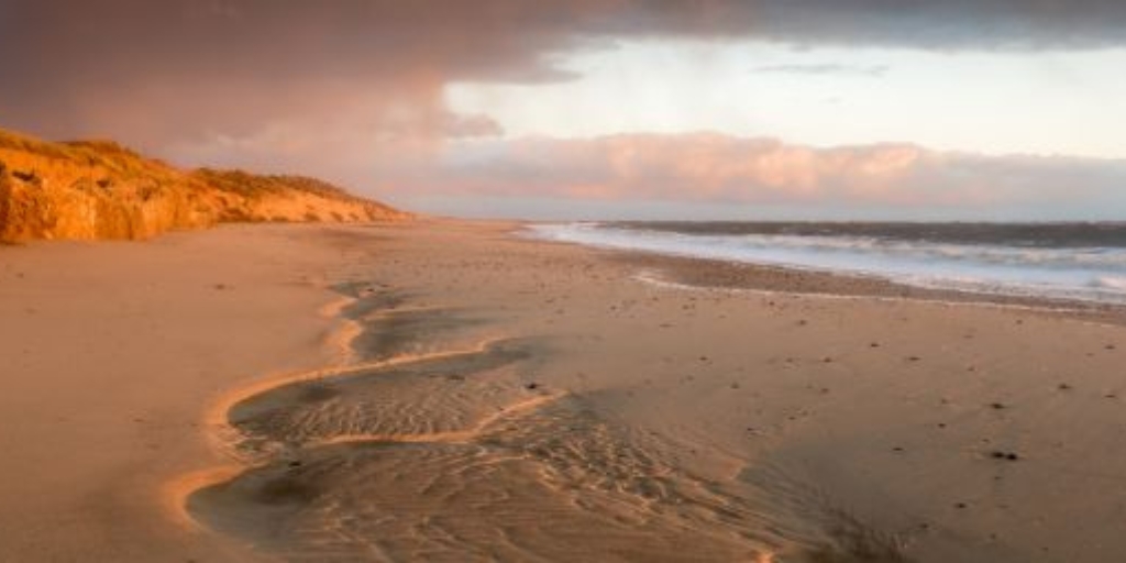 Winterton Beach in Norfolk on a sunny winter day. The sun hangs low in the sky casting an orange dusk light across the sandy dunes. The sea comes in from the right of the image, and dunes on the left. Clouds roll in from the distance.