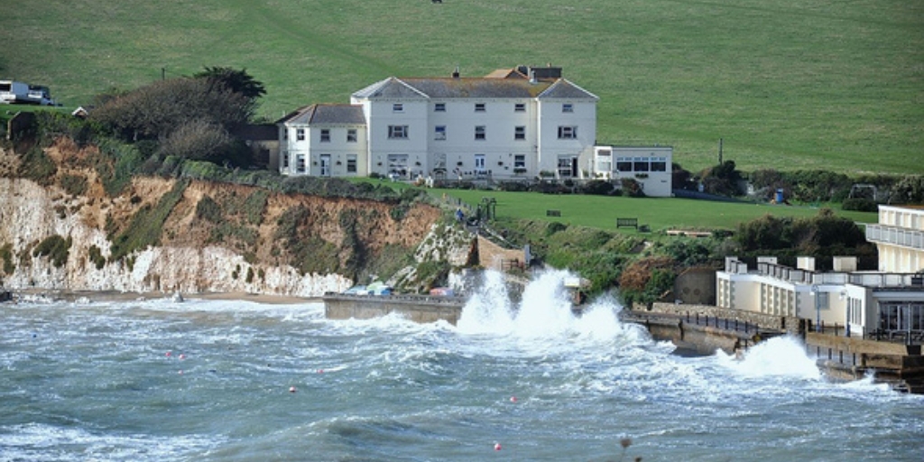 A coastal building sits atop a grassy cliff overlooking rough, choppy sea water. Waves crash forcefully against a concrete sea wall below. The pale‑coloured building stands near the edge of the eroding cliff, with wide green fields stretching out behind it. A second set of coastal buildings sits to the right, closer to the water. The scene captures a windy, energetic shoreline with dramatic waves and rugged cliffs.