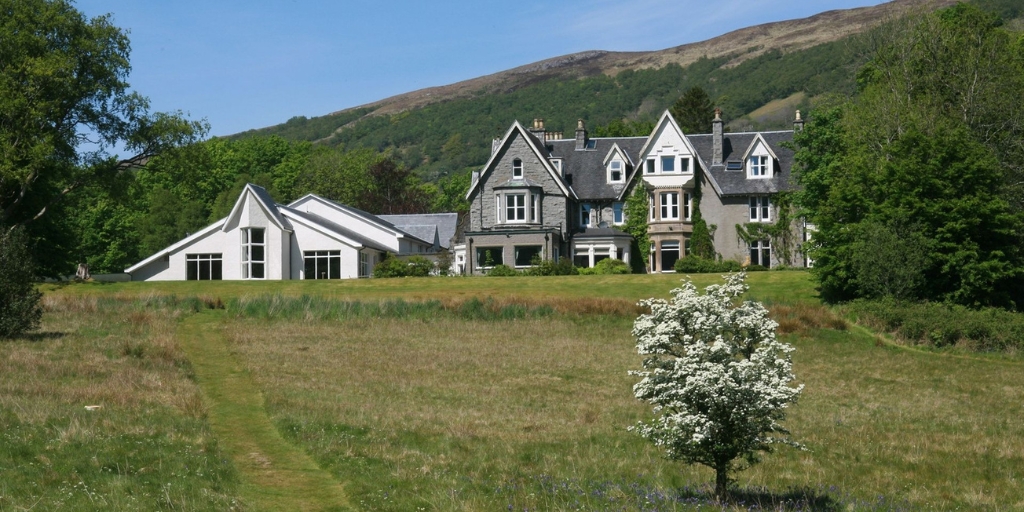 A large traditional stone house with steep gabled roofs sits at the edge of a wide grassy meadow. The building features multiple peaked dormer windows, tall chimneys, and a mix of old stone architecture alongside a modern white extension on the left. A single flowering tree stands in the foreground of the field, with wooded areas and rolling green hills rising behind the house under a clear blue sky.