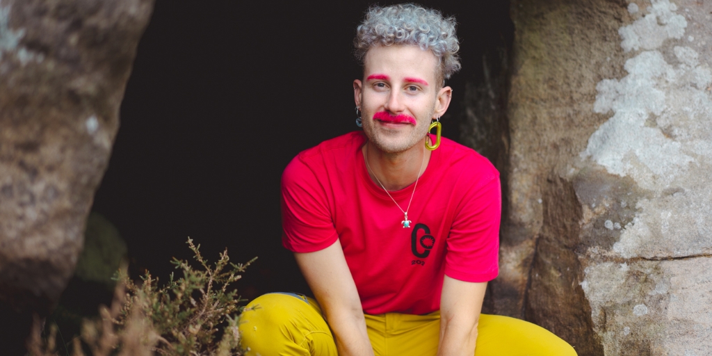 An individual is positioned outdoors between two large rock surfaces. The individual has short, curly hair and is wearing a bright red short‑sleeved shirt with a circular logo on the chest, along with yellow trousers. The individual is sitting or crouching close to the ground with hands resting forward. The person has brightly coloured cosmetic accents on the face, including vivid red coloration above each eye and a matching colour around the upper lip area. The individual is also wearing a necklace and a single, large circular earring. In the foreground, there is low vegetation, and behind the individual is a dark recessed area within the rocks, suggesting the entrance to a small cave or sheltered space.