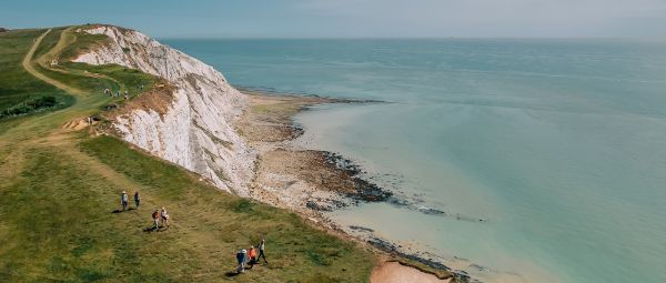 A wide aerial‑style view of a coastal cliff path. Several individuals are walking along a grassy trail that follows the edge of white chalk cliffs above a calm, pale‑blue sea. The shoreline below shows exposed rocks and shallow water. The landscape is open and expansive, with the path winding across the top of the cliffs and disappearing into the distance. The overall scene captures a large stretch of coastline from a high vantage point.