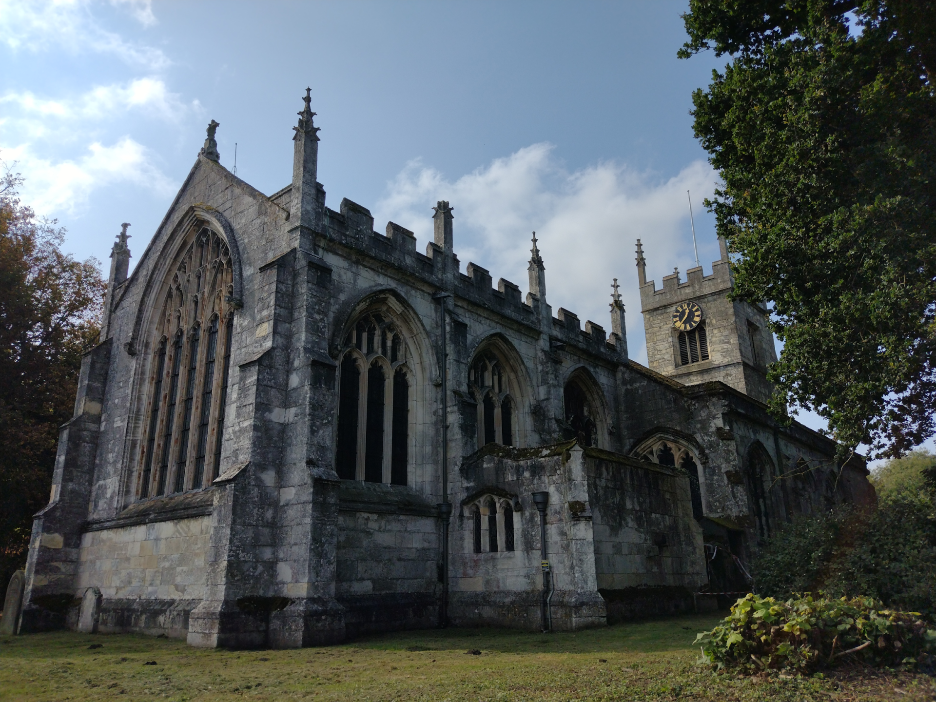 The image is of a large stone church, with a cloudy sky in the background. Surrounding the building are trees, grass, and other plants.