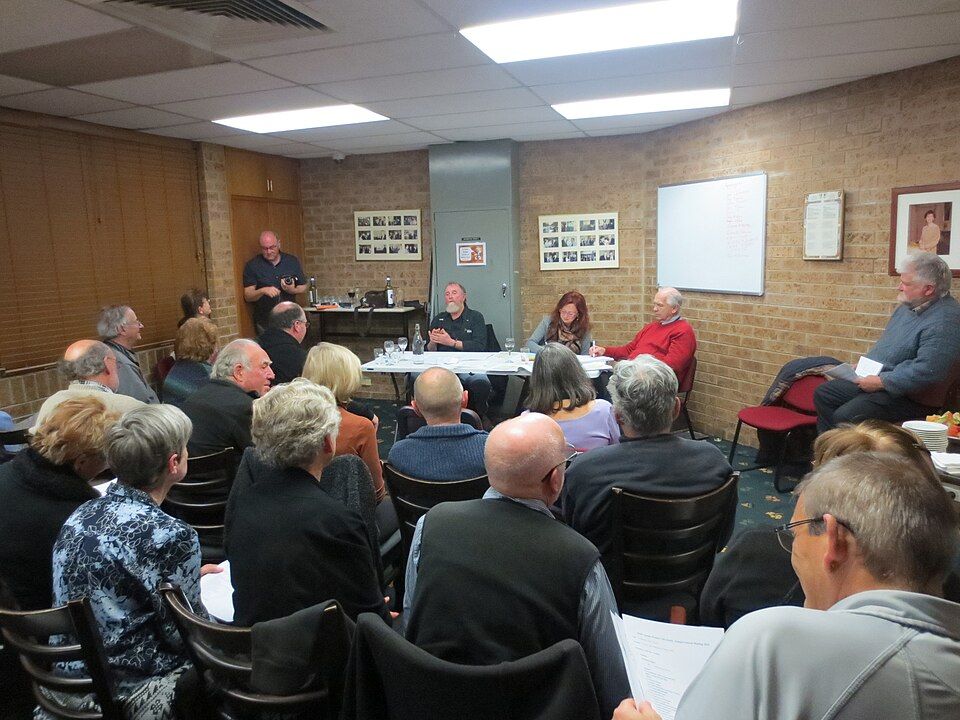 Generic photo of an AGM of a voluntary organisation showing a room full of people seated in front of a top table where committee officers are presenting. Photo is licensed via Creative Commons CC-BY-SA 4.0 by Johnscotausshared courtesy of  