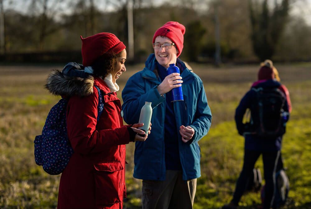 Woman and man wearing winter walking clothing,  talking and smiling while holding reusable water bottles. 