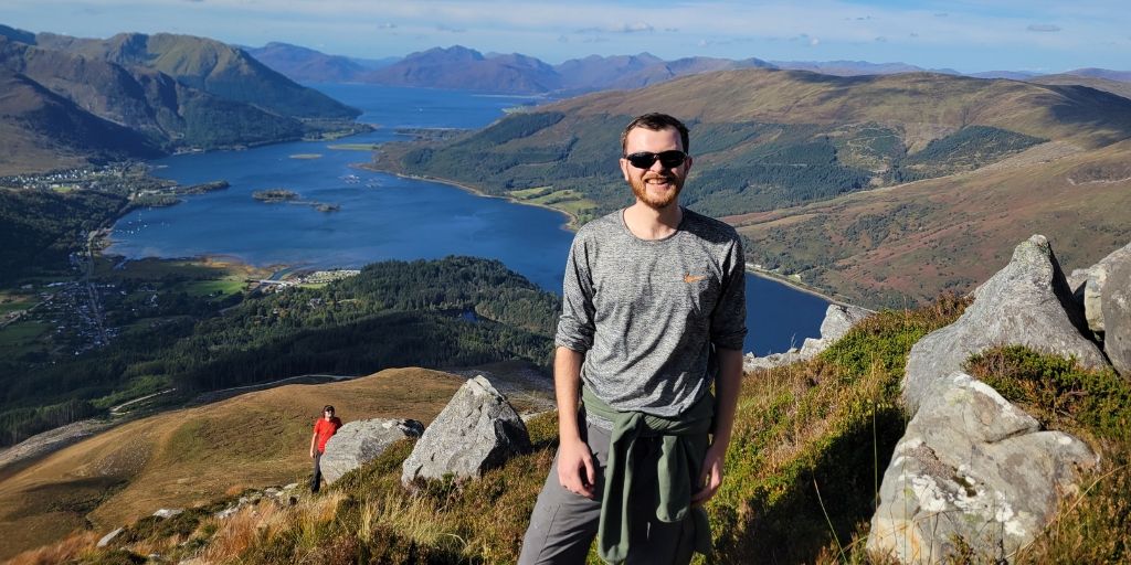 Photo of Nick stood at the top of a sunny valley, he is smiling to camera