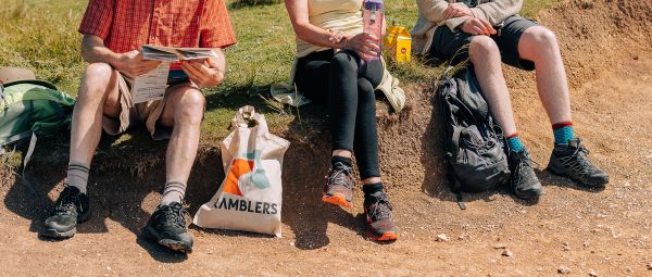 A group of three ramblers sit on the verge of a field, their legs dangling over the edge to a sandy path below, while taking a break on a summer's day. 