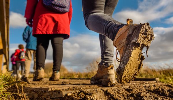 The muddy sole of a raised walking boot fills the foreground as a line of four ramblers stretches off away from the camera