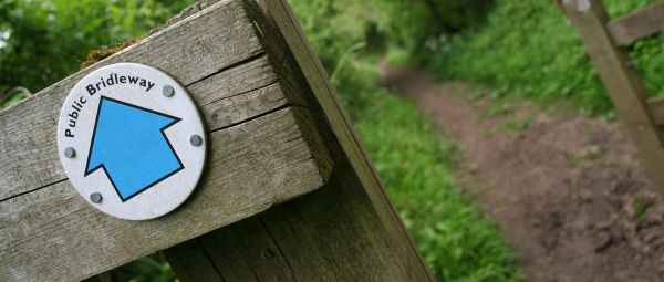 An arrow nailed to a rustic wooden gatepost indicates a public bridleway in Worcestershire, UK. Focus is firmly on the sign, with the tree-lined, horse-trodden path leading into the distance.