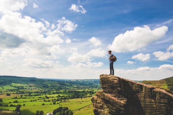 A walker looks out on green fields from a rocky outcrop in front of a blue sky and white clouds