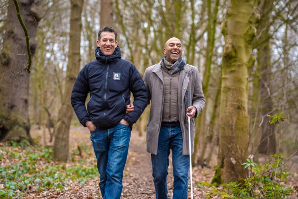 Amar Latif is holding his white cane and being guided by a sighted companion, through woodland.  Both people are laughing.