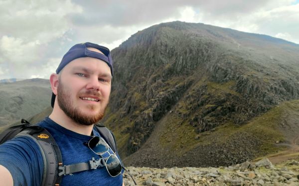 A selfie photo of Arron smiling.  There are large rugged hills behind him.