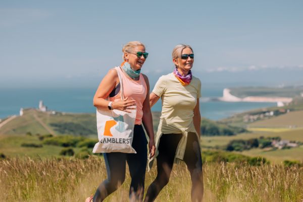 two ramblers smiling on a coastal walk with one carrying a Ramblers bag