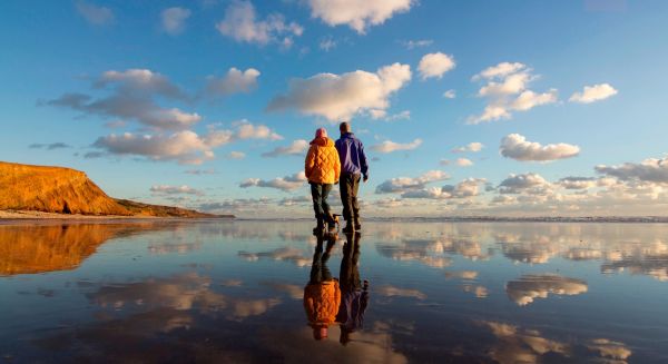 Two walkers strolling across a beach with a dog in tow.