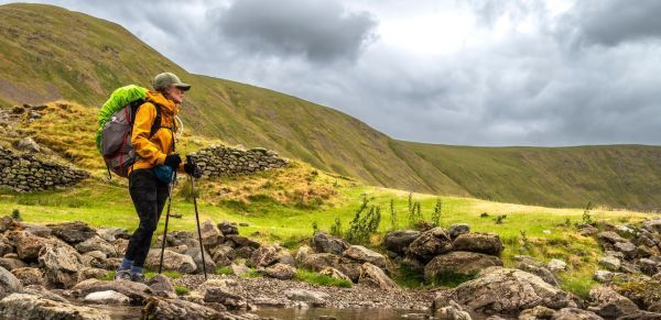 A walker with large backpack, yellow rain jacket and walking poles surrounded by large hills.