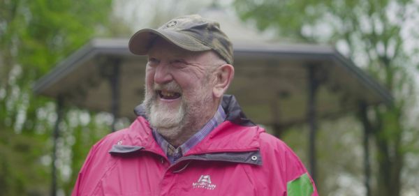 An older man with a brown cap, white beard and red waterproof laughs as he looks off camera