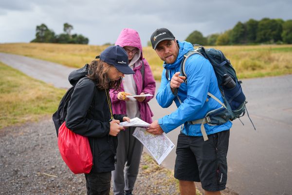 An Out There Award instructor wearing a Ramblers cap points ahead while two young people stand beside him on a path, holding maps and checking their route in open countryside.