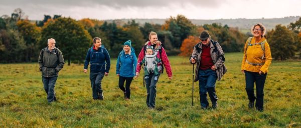 A group of ramblers walk and talk together across a lush green field surrounded by British countryside. They are in a horizontal formation and the woman in the middle is wearing a baby in a carrier