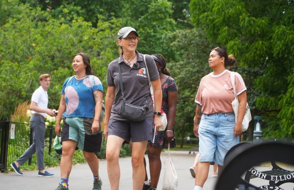 A group of people walk together along a paved path in a park surrounded by dense green trees and plants. The people wear casual clothing such as shorts, T‑shirts, and caps, and some carry bags. A person in the background walks in the opposite direction near a black metal fence. The scene is bright and outdoors, with lush greenery filling the background
