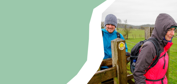 Two individuals are walking through a wooden gate in a rural field. The individual in the foreground is wearing a hooded waterproof jacket and carrying a backpack, stepping forward past a wooden post marked with a yellow circular waymarker. Behind this individual, another individual wearing a knitted hat and outdoor clothing is following through the gate. The background shows open green fields, scattered trees, and an overcast sky. 