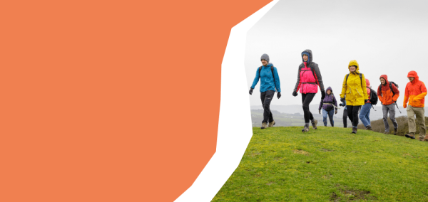 A group of individuals is walking together across a grassy hilltop in outdoor clothing. The individuals are spaced in a loose line, moving from right to left on the image. They are dressed in brightly coloured waterproof jackets, including yellow, pink, blue, orange and red, and many are carrying backpacks or wearing hoods due to the wet weather. The ground is covered in short green grass, and the sky is overcast with grey cloud. 