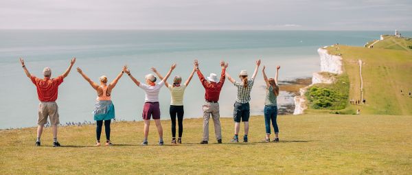 A group of seven individuals stands on a grassy cliff top overlooking the sea. They are shown from behind, standing side by side in a row with arms raised in the air. The cliff edge drops down to the shoreline below, where white chalk cliffs meet turquoise water. The sky is bright and lightly clouded, creating a clear coastal view. The group is positioned near the centre of the image with the coastline curving away into the distance on the right.