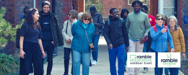 A group of people is walking together outdoors along a paved pathway between two brick buildings. The group is arranged loosely in a line, moving toward the camera. Several individuals are wearing outdoor jackets, while others wear lighter clothing such as T‑shirts or sweatshirts. Some carry backpacks or shoulder bags. The path is surrounded by planted greenery, including shrubs and small trees. 