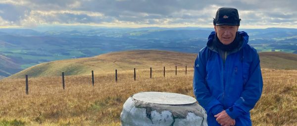 Colin on top of a hill, standing next to an interpretative plaque in 2021.