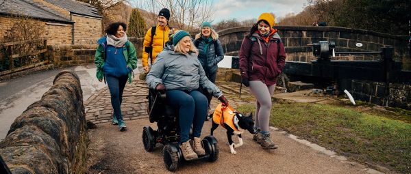 A group of ramblers, one using a motorised wheelchair, walk along a canal towpath by a lock