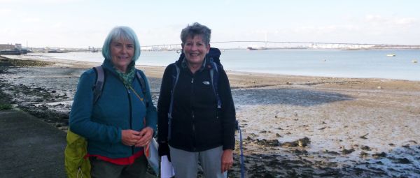 Two women standing by a river shore, in front of a large bridge