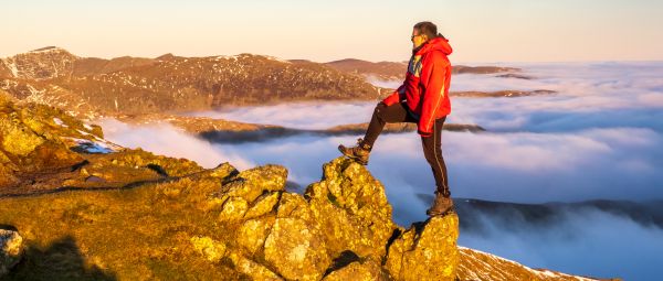 A man stands on top of Helvellyn, looking out over the clouds below hi, with one foot each on two adjacent rocks