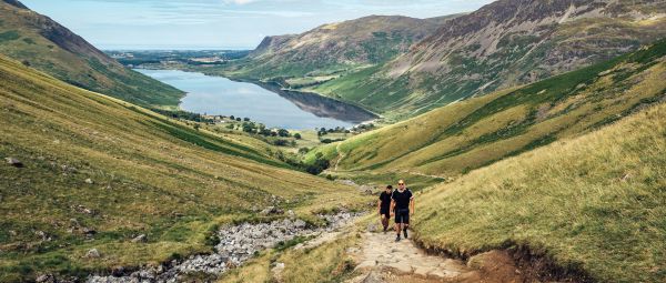 Two ramblers approach the camera, climbing a paved path with a shimmering blue lake in the background below them