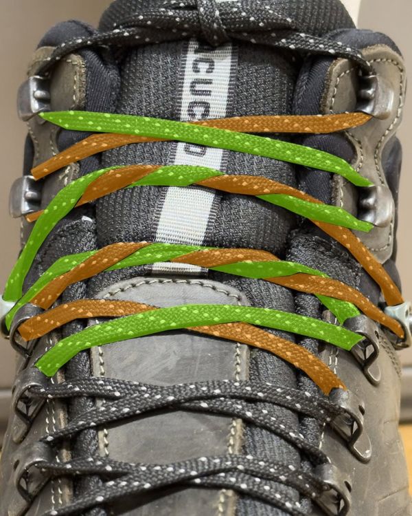Close-up of the upper section of a grey hiking boot. The laces alternate in green and orange to highlight the steps. The laces cross over each other in a standard criss-cross pattern, but at two points the crossovers are doubled, forming a surgeon’s knot that locks the tension in place. The surgeon’s knots sit at the pressure points of the foot, keeping the lacing snug and preventing loosening during movement.