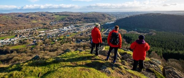 Three people in red jackets, standing on top of a hill, looking down at the valley and town below