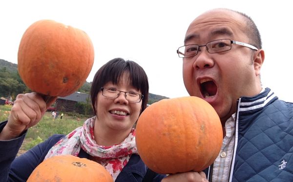 A woman and man holding up pumpkins, as though to take a bite from one